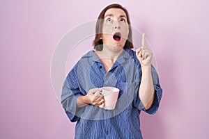 Middle age hispanic woman drinking a cup coffee amazed and surprised looking up and pointing with fingers and raised arms