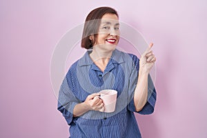 Middle age hispanic woman drinking a cup coffee with a big smile on face, pointing with hand finger to the side looking at the