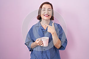 Middle age hispanic woman drinking a cup coffee cheerful with a smile on face pointing with hand and finger up to the side with