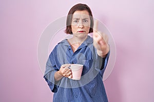 Middle age hispanic woman drinking a cup coffee pointing with finger up and angry expression, showing no gesture