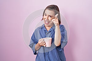 Middle age hispanic woman drinking a cup coffee pointing fingers to camera with happy and funny face