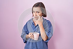 Middle age hispanic woman drinking a cup coffee pointing to the eye watching you gesture, suspicious expression