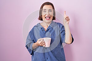 Middle age hispanic woman drinking a cup coffee smiling amazed and surprised and pointing up with fingers and raised arms