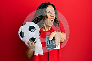 Middle age hispanic woman football hooligan holding ball using tv control angry and mad screaming frustrated and furious, shouting