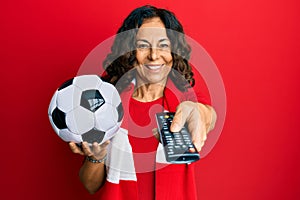 Middle age hispanic woman football hooligan holding ball using tv control smiling with a happy and cool smile on face