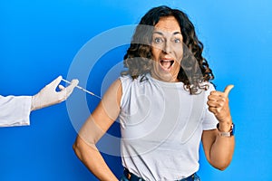 Middle age hispanic woman getting vaccine pointing thumb up to the side smiling happy with open mouth