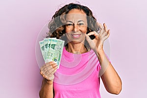 Middle age hispanic woman holding 10000 south korean won banknotes doing ok sign with fingers, smiling friendly gesturing