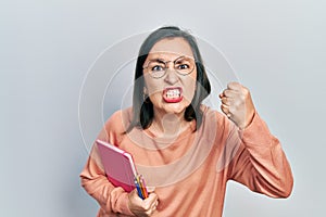 Middle age hispanic woman holding book and color pencils annoyed and frustrated shouting with anger, yelling crazy with anger and