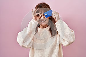 Middle age hispanic woman holding credit card covering eye smiling happy doing ok sign with hand on eye looking through fingers