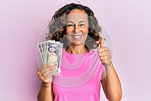 Middle age hispanic woman holding japanese yen banknotes smiling happy and positive, thumb up doing excellent and approval sign