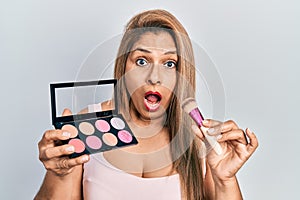 Middle age hispanic woman holding makeup brush and blush afraid and shocked with surprise and amazed expression, fear and excited