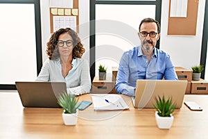 Middle age hispanic woman and man sitting with laptop at the office relaxed with serious expression on face