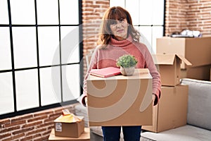 Middle age hispanic woman moving to a new home holding cardboard box clueless and confused expression