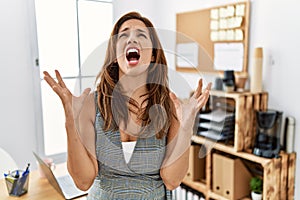 Middle age hispanic woman at the office crazy and mad shouting and yelling with aggressive expression and arms raised