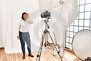 Middle age hispanic woman posing as model at photography studio smiling happy doing ok sign with hand on eye looking through