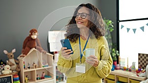 Middle age hispanic woman preschool teacher using smartphone drinking coffee at kindergarten