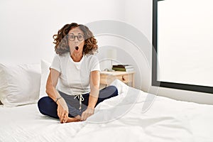 Middle age hispanic woman sitting on the bed at home afraid and shocked with surprise expression, fear and excited face