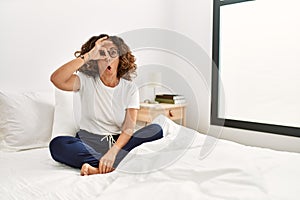 Middle age hispanic woman sitting on the bed at home doing ok gesture shocked with surprised face, eye looking through fingers