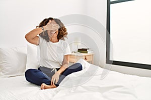 Middle age hispanic woman sitting on the bed at home smiling and laughing with hand on face covering eyes for surprise