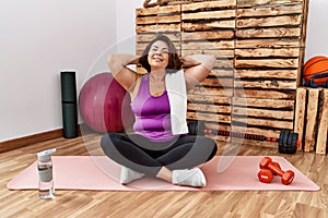 Middle age hispanic woman sitting on training mat at the gym relaxing and stretching, arms and hands behind head and neck smiling
