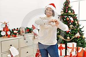 Middle age hispanic woman standing by christmas tree smiling in love doing heart symbol shape with hands