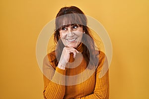 Middle age hispanic woman standing over yellow background with hand on chin thinking about question, pensive expression