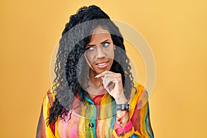 Middle age hispanic woman standing over yellow background with hand on chin thinking about question, pensive expression