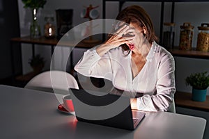 Middle age hispanic woman using laptop at home at night peeking in shock covering face and eyes with hand, looking through fingers