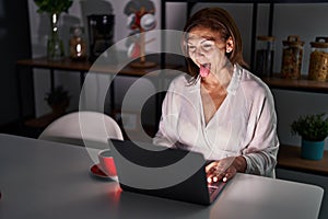 Middle age hispanic woman using laptop at home at night sticking tongue out happy with funny expression