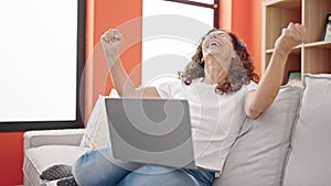 Middle age hispanic woman using laptop sitting on sofa with winner expression at home