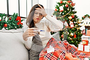 Middle age hispanic woman using thermometer sitting by christmas tree at home