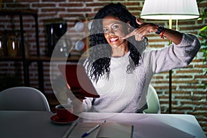 Middle age hispanic woman using touchpad sitting on the table at night smiling cheerful showing and pointing with fingers teeth