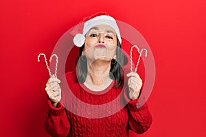 Middle age hispanic woman wearing christmas hat holding candy looking at the camera blowing a kiss being lovely and sexy