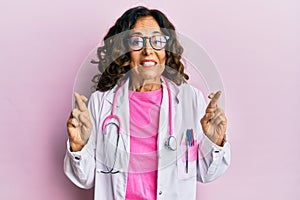 Middle age hispanic woman wearing doctor uniform and glasses gesturing finger crossed smiling with hope and eyes closed