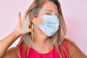 Middle age hispanic woman wearing medical mask smiling with hand over ear listening and hearing to rumor or gossip