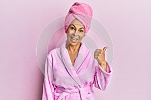 Middle age hispanic woman wearing shower towel cap and bathrobe smiling with happy face looking and pointing to the side with