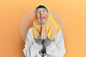 Middle age hispanic woman wearing traditional islamic hijab scarf begging and praying with hands together with hope expression on