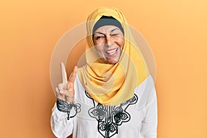 Middle age hispanic woman wearing traditional islamic hijab scarf smiling with happy face winking at the camera doing victory sign