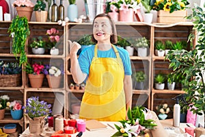 Middle age hispanic woman working at florist shop pointing thumb up to the side smiling happy with open mouth