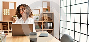 Middle age hispanic woman working at the office wearing glasses smiling with happy face looking and pointing to the side with