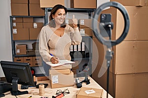 Middle age hispanic woman working at small business ecommerce doing online tutorial smiling happy and positive, thumb up doing