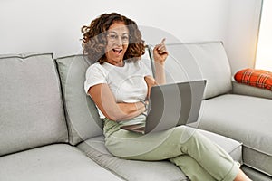 Middle age hispanic woman working using computer laptop at home smiling happy pointing with hand and finger to the side