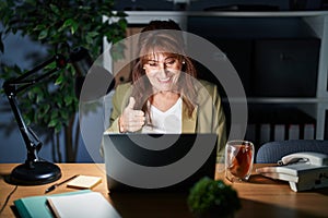 Middle age hispanic woman working using computer laptop at night doing happy thumbs up gesture with hand