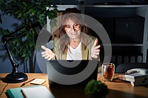 Middle age hispanic woman working using computer laptop at night looking at the camera smiling with open arms for hug