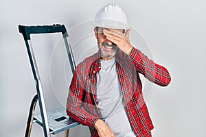 Middle age hispanic worker man using ladder stressed and frustrated with hand on head, surprised and angry face