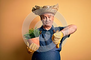 Middle age hoary farmer man wearing apron and hat holding plant pot over yellow background with angry face, negative sign showing