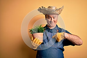 Middle age hoary farmer man wearing apron and hat holding plant pot over yellow background with surprise face pointing finger to