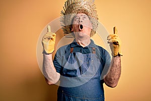 Middle age hoary farmer man wearing apron and hat over isolated yellow background amazed and surprised looking up and pointing