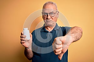 Middle age hoary man holding medicine pills standing over isolated yellow background with angry face, negative sign showing