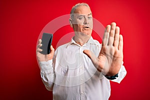 Middle age hoary man holding smartphone showing screen over isolated red background with open hand doing stop sign with serious